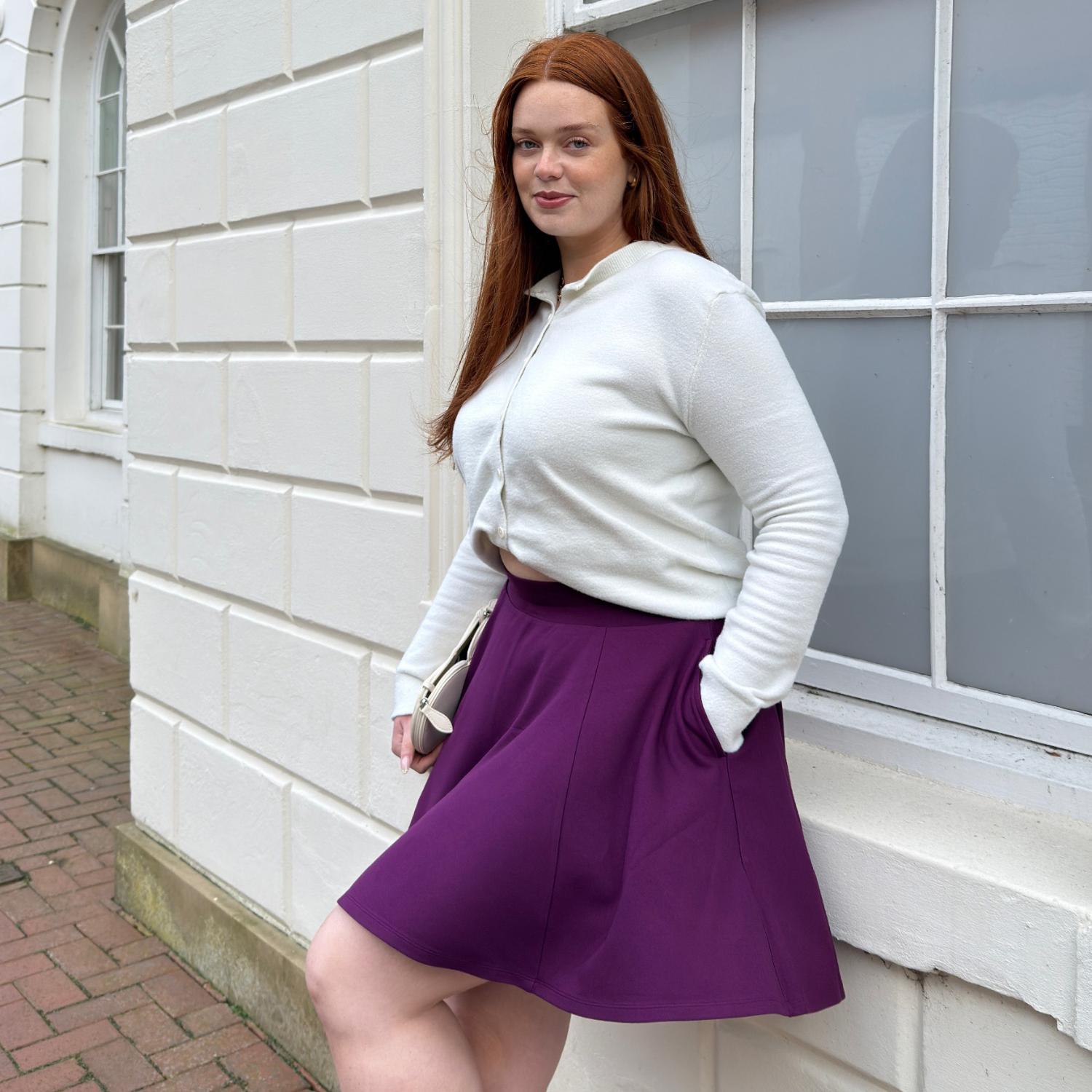 Woman in a white shirt and purple skirt standing against a white wall.