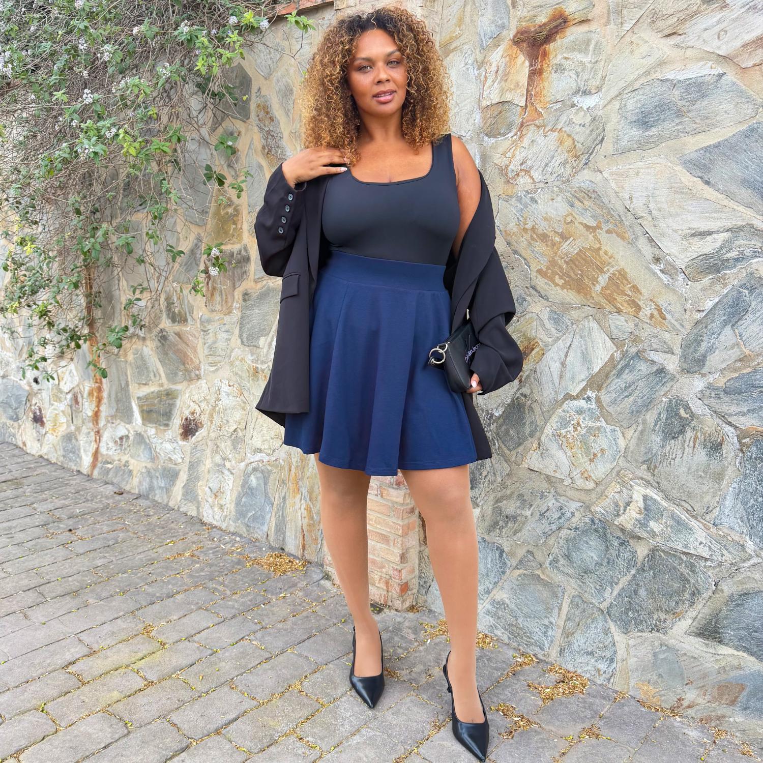 Woman in a navy dress and black jacket standing against a stone wall.