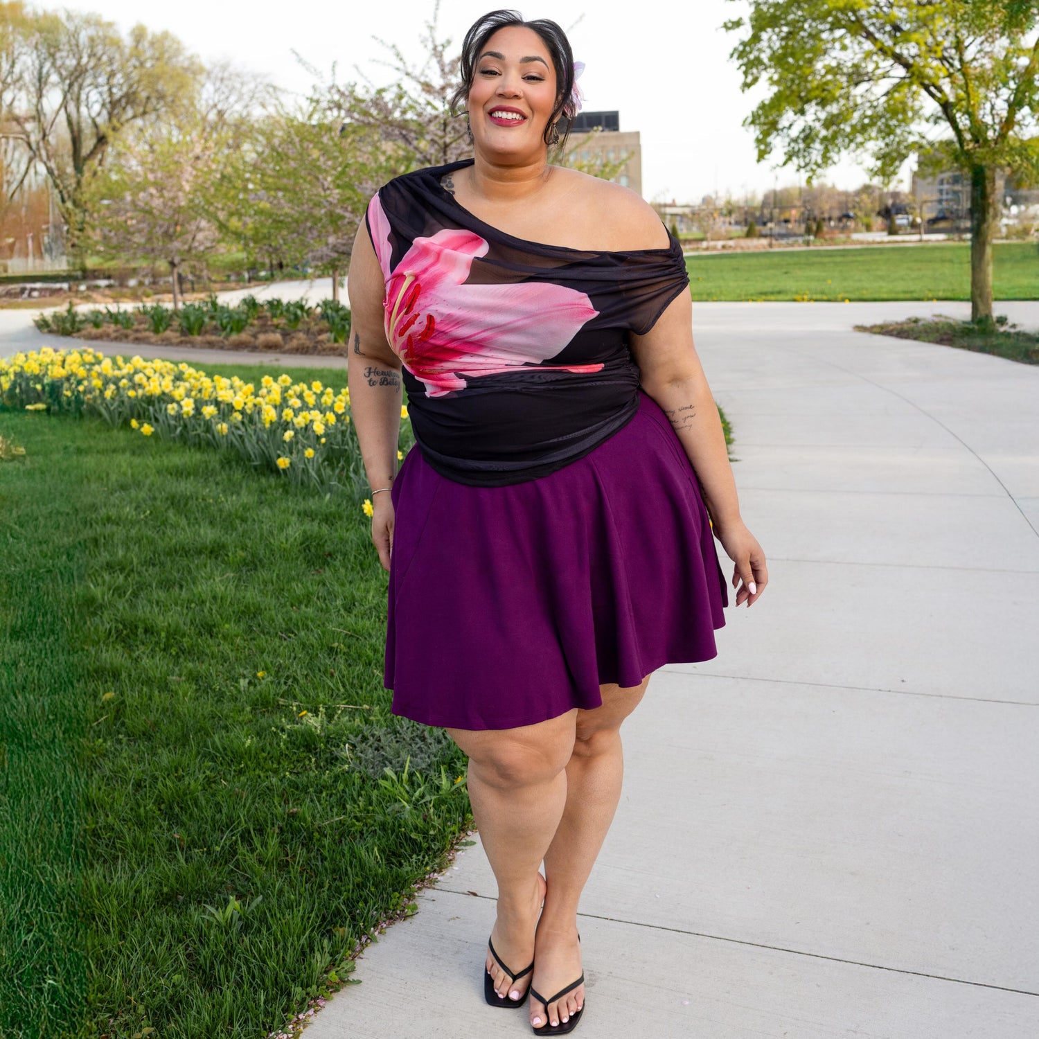 Woman wearing a floral one-shoulder top and purple skirt standing outdoors with greenery and flowers in the background.