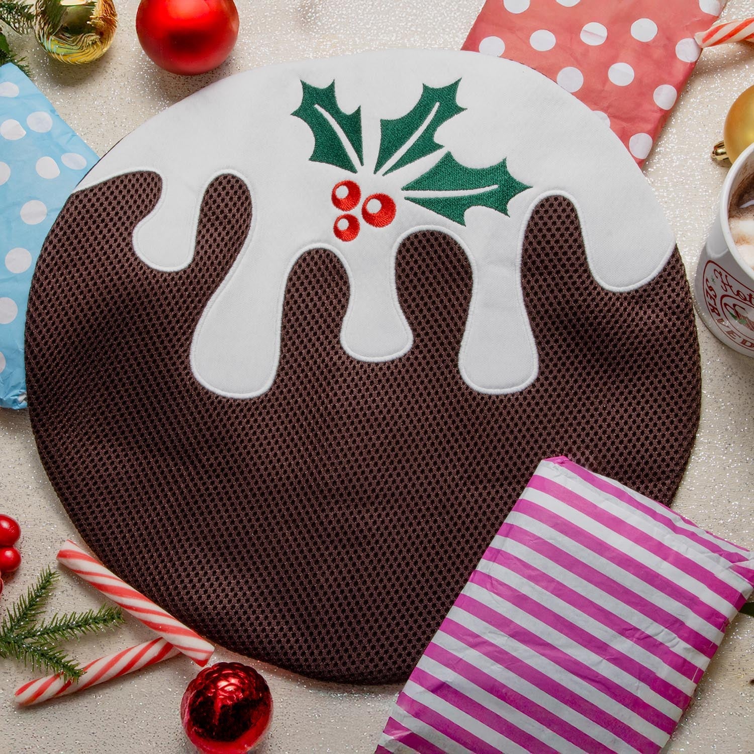 A festive flat lay photo for the Snagvent Advent Calendar 2025, centered on a large, round washbag designed to look like a Christmas pudding. The bag features a brown mesh body, white fabric 'icing' dripping down the top, and embroidered green holly leaves with red berries. It is placed on a glittery surface, surrounded by Christmas items including colorful paper gift bags, candy canes, red and gold baubles, and a mug of hot chocolate.